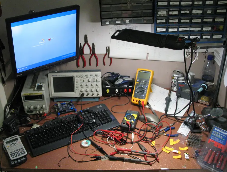 A cluttered home electronics hobbyist workbench with tools, instruments, and component storage